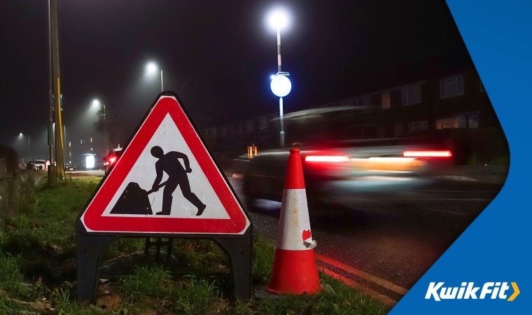 Roadworks sign on the side of a grassy road with a passing car.