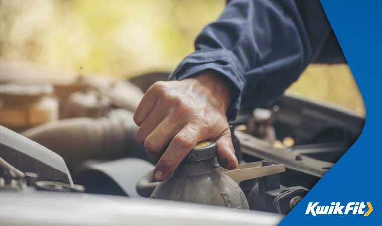 Technician wearing a boiler suit twisting the cap of the engine coolant compartment under the bonnet of a vehicle.