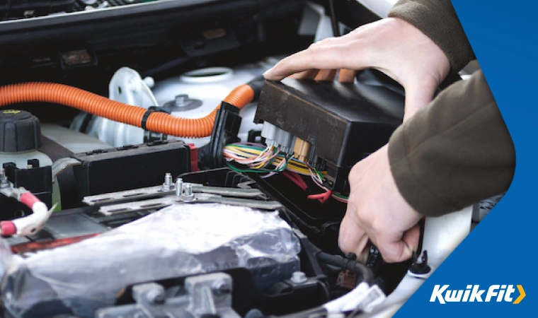 Two hands carefully lift a fuse box up from underneath the bonnet of the car. It is still attached to the car and wires are visible.