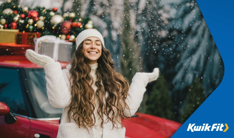 A woman stands smiling in the snow outside her car at Christmas time.